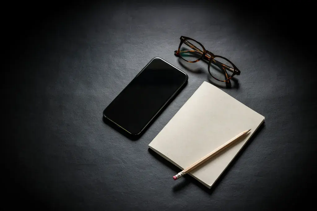 Un celular con la pantalla apagada, unos lentes y una libreta cerrada sobre una mesa oscura